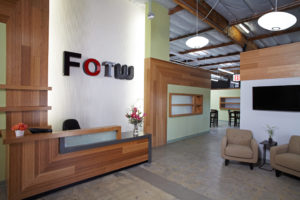 Rendered view of a modern office entry space. Includes wood wall panels against a green wall, a wooden front desk below a company logo, and two chairs below a television.