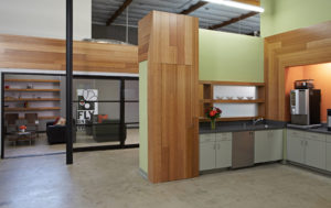 Rendered view of a modern office kitchen. Includes wood wall panels against a green wall, a kitchen with white cabinets and orange wall, and a view into a conference room through an open glass door.