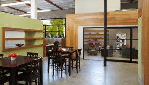 Rendered view of a modern office communal space. Includes wood wall panels against a green wall, multiple counter-height tables, and a view into a conference room through an open glass door.