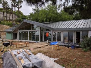 Photo of construction taking place on the front facade of a modern house. The foreground features a few wooden planks and furniture pieces on the floor, while the background shows a man on a ladder facing a window.