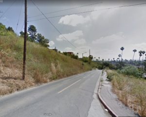 Google Maps screenshot of a hillside from the street. A road runs through the middle of the image, with dry grass on either side and views of a suburban neighborhood in the background.