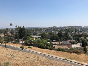 Photo from a hilltop overlooking a suburban neighborhood. The foreground includes dry grass and a road stretching across the image, while the background features multiple houses and trees.