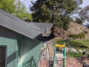 Photo of the construction of a modern house roof. Multiple ladders are shown around the black roof and green walls in the foreground, while the background features a dirt hillside with a few trees.