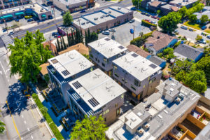 Aerial photo of eight modern townhouses and the surrounding neighborhood. Shows the rooftop outdoor spaces, driveway, and the ground floor patio bordering the sidewalk.