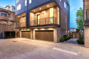 Photo of the interior courtyard serving multiple modern townhouses. Shows the driveway, pedestrian entrance, garage doors, and balconies above the garages.