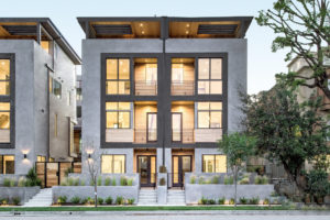 Photo of the front facade for a modern townhouse. Shows the entry door, multiple balconies, rooftop space, and concrete and wood exterior walls.