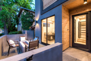 Photo of a front patio for a modern townhouse. Shows the concrete and wood exterior facade, the front entryway, a concrete fire pit, and a glass window looking into the townhouse.