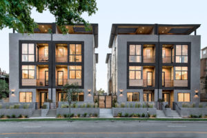 Photo of the front facade for four modern townhouses. Shows the entry doors, multiple balconies, multiple rooftop spaces, and concrete and wood exterior walls.