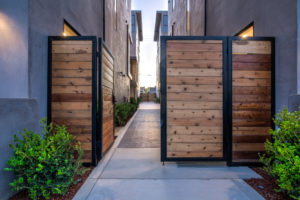 Photo of an entry gate leading into a complex of multiple modern townhouses. The wooden gate is left open to show the driveway, with the concrete walls of the townhouses flanking either side.