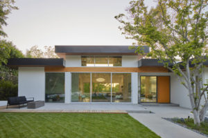 Rendering of the backyard and back facade of a modern house. Includes a concrete patio and walkway with an outdoor sofa, a floor to ceiling glass sliding door going into the living room, and a grass yard.