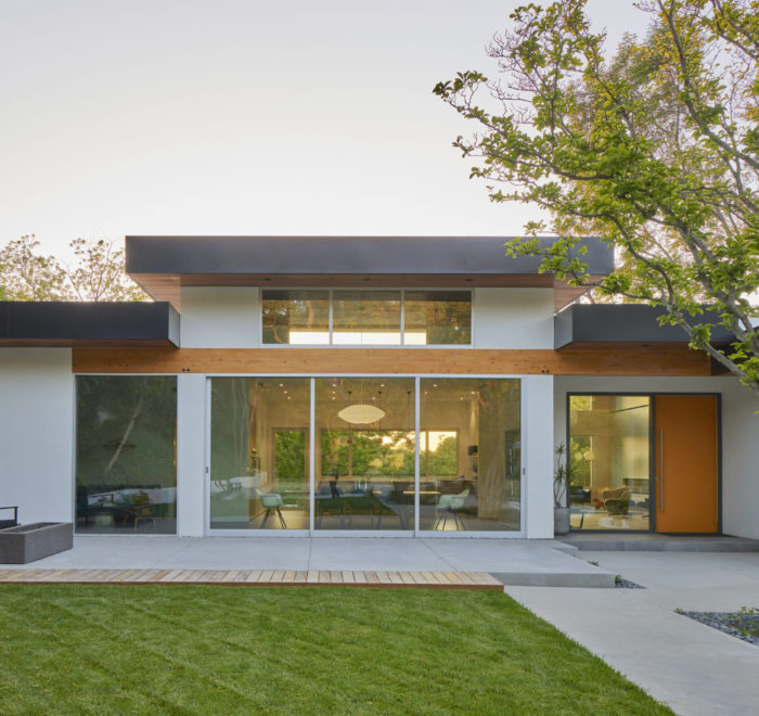 Rendering of the backyard and back facade of a modern house. Includes a concrete patio and walkway with an outdoor sofa, a floor to ceiling glass sliding door going into the living room, and a grass yard.