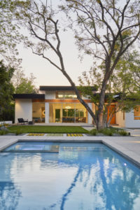 Rendering of the backyard and facade of a modern house. Includes a concrete patio and walkway, a floor to ceiling glass sliding door going into the living room, and a grass yard with a pool.