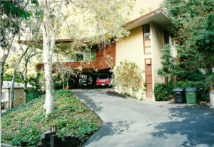 Photo of the parking lot and lower entry of an older house. Features a stone driveway, a wood and white stucco facade, and a pair of trees in the foreground.