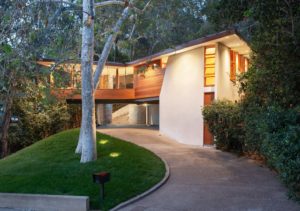 Photo of a parking lot and lower entry of a modern house. Features a stone driveway, a wood and white stucco facade, and a pair of trees placed in the front lawn.