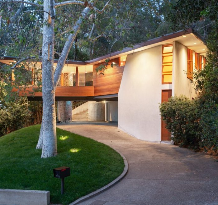 Photo of a parking lot and lower entry of a modern house. Features a stone driveway, a wood and white stucco facade, and a pair of trees placed in the front lawn.