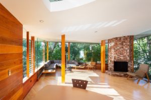 Photo of a living room within a modern house. Features a white stucco ceiling, wooden walls and ceiling, a red brick hearth and fireplace, and glass windows running around the room.