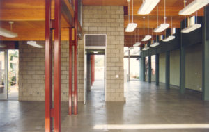 View of Office Interior before construction. Includes multiple hanging lights, a wooden roof, a brick wall, and both red and blue steel columns.