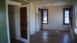 Photo of an empty run down building. Includes green and white walls, a wood floor, black framed windows, and an air conditioner unit towards the back of the unit.