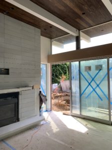 Photo showing construction of a modern house. The interior features a wooden ceiling and gray brick wall fireplace, while the glass door has a "X" of blue tape on it and reveals a trench in the backyard.