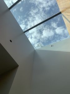 Photo pointed upwards at a skylight within a modern house. The sunlight shines against both a white plaster wall and a wooden wall.