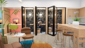 Renderings of a modern office's private call room located between the primary workspace and kitchen area. Each room features a blue geometrical wallpaper, a wooden table and cushioned chair mounted to the wall, and a hanging overhead light.