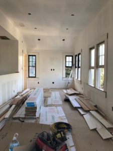 Construction photo showing work on the upstairs of a modern house. The foreground features lumber, floor covering, and equipment laying on the ground, while the background features five open windows.