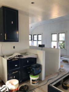 Construction photo showing work on the upstairs of a modern house. The foreground features a partially built black cabinet and equipment laying on the ground, while the background features five open windows.