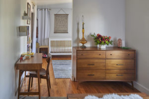 Photo of nursery partially hidden behind a wall. The modern house includes white plaster walls and wood floors, a wooden desk, and a baby's crib at the back of the photo.