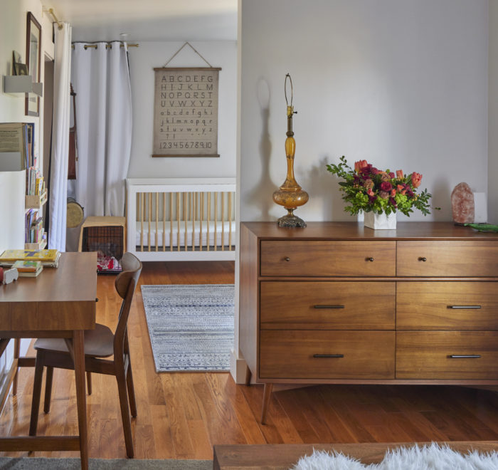 Photo of nursery partially hidden behind a wall. The modern house includes white plaster walls and wood floors, a wooden desk, and a baby's crib at the back of the photo.