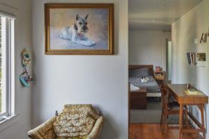 Photo of bedroom partially hidden behind a wall. The modern house includes white plaster walls and wood floors, a wooden desk, and a photo of a dog prominently featured on the wall hiding the bed.