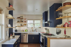 Photo showing the kitchen of a modern home. Includes blue cabinets with white marble countertops in the kitchen a gas stove and dishwasher,, and wooden shelves holding various items.