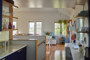 Photo showing the kitchen and living room of a modern home. Includes colorful pillows adorning the dining table seating, blue cabinets with white marble countertops in the kitchen, and wooden shelves holding various items.