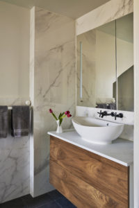 Photo of a bathroom within a modern house. Includes both white marble and plaster walls, a mirror with a built in light, and a wooden counter with a marble countertop.