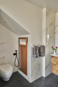 Photo of a bathroom within a modern house. Includes both white marble and plaster walls, a wooden cabinet by the toilet, and a wooden counter with a marble countertop.