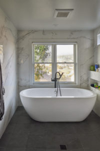 Photo of a bathroom within a modern house. The open concept bathroom has marble walls that have built-in shelves, black tile floors, and a large bathtub.