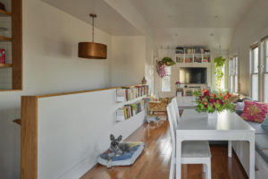 Photo of main living space within a modern house. Includes a white dining table with chairs and a cushioned bench, a living room with a wall-mounted television in the back, and a staircase to the garage on the left side.