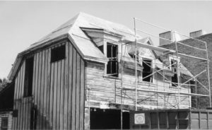 Black-and-white photo of construction work on a modern house. Scaffolding stands against the building on the front facade, and wood is also laying on the roof.