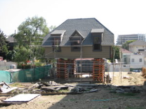 Photo of construction work on the second floor of a modern house. With no first floor present, wooden turning blanks are stacked to hoist the second floor of the building above the ground. There is excess wood laying in the foreground of the photo, and scaffolding surrounds the house.