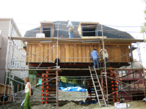 Photo of construction work on the second floor of a modern house. With no first floor present, wooden turning blanks are stacked to hoist the second floor of the building above the ground