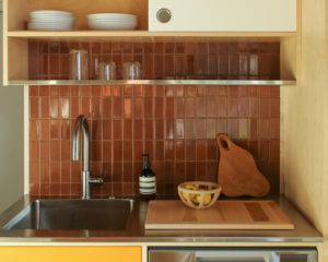 A stainless steel countertop with a sink and dishwasher bordering a brown tile wall. A metal wire shelf and wooden cabinetry lie overhead and to the right side.