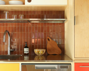 A stainless steel countertop with a sink and dishwasher placed against a brown tile wall. A metal wire shelf and wooden cabinetry lie overhead and to the right side.