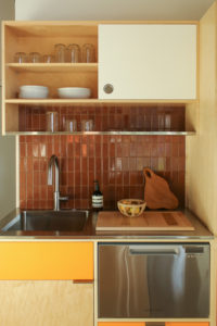 A stainless steel countertop with a sink and dishwasher bordering a brown tile wall. A metal wire shelf and wooden cabinetry lie overhead and to the right side.