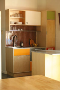 A kitchen featuring a counter space with an sink and dishwasher against a brown tile wall. A metal wire shelf and colored wooden cabinetry lie overhead and to the right side.