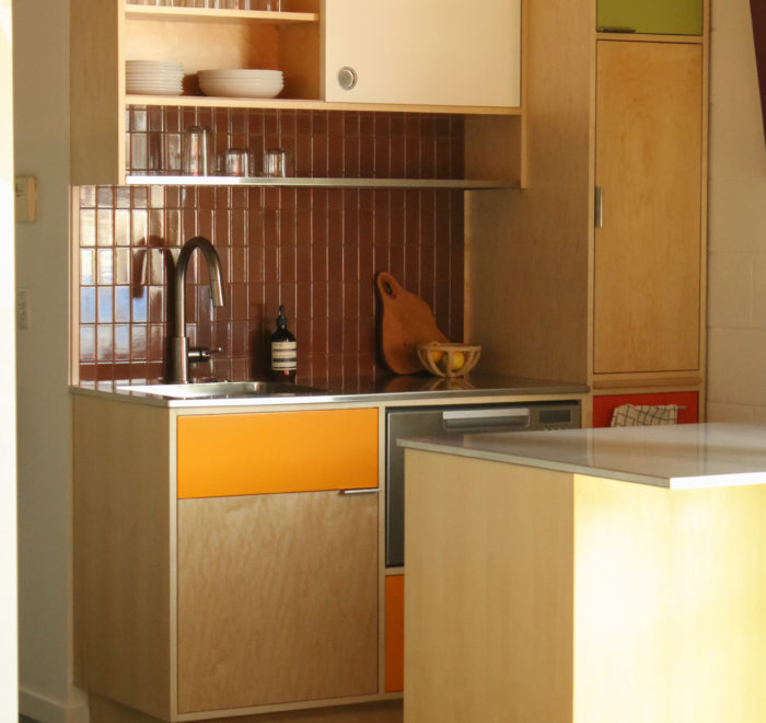A kitchen featuring a counter space with an sink and dishwasher against a brown tile wall. A metal wire shelf and colored wooden cabinetry lie overhead and to the right side.